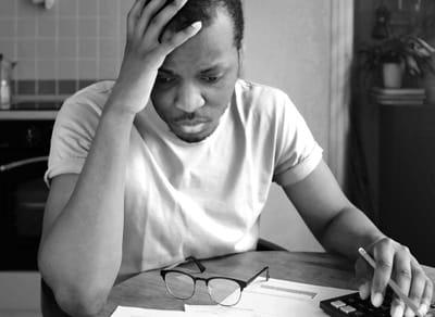 一个黑人坐在餐桌旁，双手托着头，看着账单和文件